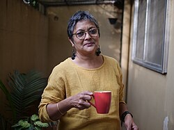 Mamta Sagar wearing a yellow sweater and glasses, holding a red mug in one hand, smiling at camera