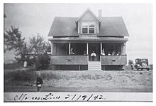 Une photographie en noir et blanc d'une maison à un étage et demie, avec une galerie en avant sur laquelle se tiennent 7 personnes. À droite de la maison, une voiture sombre est stationnée. En avant plan, un enfant aux cheveux clairs se tient sur la route. À gauche de la maison, un bosquet.