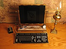 a flat screen monitor of unusual riveted edging with a keyboard containing raised keys as found on mechanic typewriters. The pair accompanied by a Victorian gas lamp.