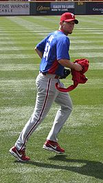A man in grey pants, a blue baseball jersey, and a red baseball cap with "P" on it jogs on a grass field while carrying a red article of clothing in his right hand.