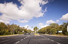 Foto einer vierspurigen Straße mit Ampel, an der auf der linken Seite gerade Autos halten. Links und rechts der Straße sieht man jenseits der Ampel Bushaltestellen mit Wartehäuschen.