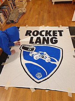 Person som målar en tvåpinnsflagga på golvet i ett rum. På flaggan står det ROCKET LANG och är en bild på bilen Octane från spelet Rocket League där den nickar en puck.