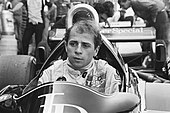 A black and white photograph of a man sitting inside a stationary racing car without a racing helmet but in racing overalls