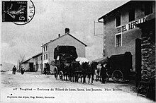 Carte postale noir et blanc. Un chemin de terre est bordé d'un côté de cinq maisons à un ou deux étages. L'un d'eux, à deux étages, porte l'enseigne d'un hôtel. Devant stationnent deux diligences.