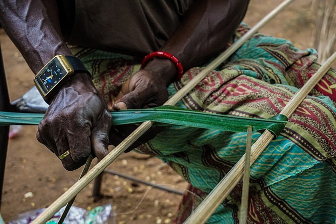 He is making a new roof for his shop, he tells me. He wants me to make pictures. There are palm leaves all over, some tied up in a bunch, some lying underneath the sun, others thatched already and staked up in a pile. On his hands are two long,thinly carved sticks that would function as the skeletal frame for a pair of thatch.