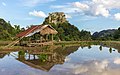 Reflet dans l'eau de montagnes, d'un abri en bois et de gerbes de riz vert éparpillées dans une rizière, avec nuages et ciel bleu, un jour ensoleillé durant la mousson, dans la campagne de Vang Vieng.