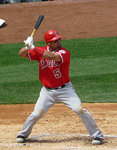 A brown-skinned man in a red baseball jersey and while pants takes a right-handed baseball batting stance.