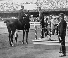 Photo noir et blanc. Le roi, en grand uniforme, de profil sur une estrade, tend la médaille à Cariou, qui est à cheval, vu de dos.