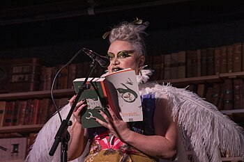 A dashingly made up women with short blond curly hair is behind a microphone reading from her book. Her outfit includes feathered wings.