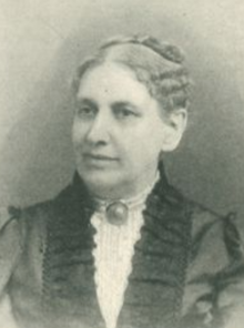 An older white woman, greying hair parted center and dressed to the crown, wearing a high-collared white lacy blouse iwth a pin at the throat, and a dark overdress with a ruffled neckline