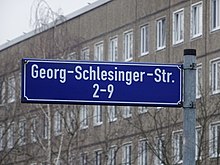 Blaues Straßenschild mit weißer Schrift. Aufschrift: Georg-Schlesinger-Str. 2–9. Im Hintergrund sind kahle Bäume vor einem Plattenbau zu sehen