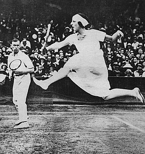 Lenglen hitting her signature leaping volley shot at the net from the advantage side of the court, with her partner Gerald Patterson watching also at the net on the deuce side of the court