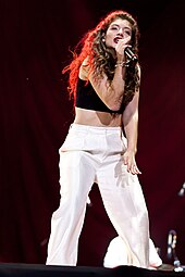 Lorde performs on-stage in a black crop top and white pants under a red spotlight.