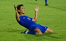 Chhetri on his knees in a blue football shirt and blue football shorts. His arms are outstretched and he is smiling after just scoring a goal.