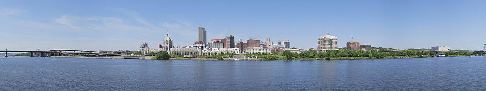 A panorama shows a river in the bottom half, crossed by a highway bridge on left; building towers are seen around the center, where a green zone on the bank of the river is seen, which extends to the right extreme of the image.