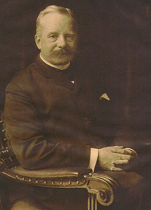 A middle aged man wearing an old style suit, is pictured sitting on chair. The old picture is black and white.