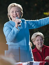 Photograph of Clinton in a light blue suit, holding a microphone and speaking in front of Elizabeth Warren who is seated behind her