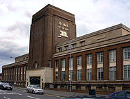 A long, tall three-storey building (with a very tall central square tower providing a fourth storey) made of brown brick.