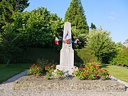 Photographie d'un monument en forme de petit obélisque au milieu d'une aire gravillonnée entourée de haies