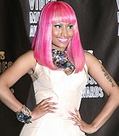 A tan-skinned woman in a bright pink wig poses with hands on both side of her hips. Smiling, she stands before a black background and has Mandarin characters tattooed on her right arm.