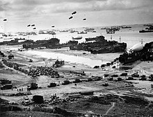 Photographie d'une plage où se trouvent des dizaines de véhicules. À l'arrière-plan, des dizaines navires de toutes tailles, dont certains sont échoués pour débarquer les équipements, sont protégés par des ballons de barrage.