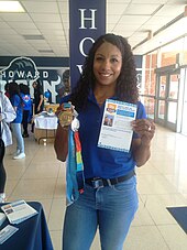Maritza Correia in blauer Bluse und Jeans hält in der rechten Hand mehrere Medaillen und in der linken Hand ein Flugblatt.