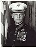 A black and white image of Pless wearing his military dress uniform with hat and medals. His Medal of Honor is visible around his neck and there is an American flag in the background.