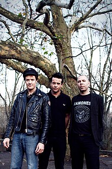 Three men stand in front of a bare tree. All are shown in three-quarter body shot and facing forward. Man at left is tallest, he has dark hair and wears a dark cap, a dark leather jacket which is unzipped, and blue jeans. He holds his arms along his sides. Middle man is slightly shorter, with dark clothes and his arms behind his back. He is partly behind and obscured by first man. Third man is shortest, his head hair is very short and sparse and his hands are behind his back. He wears a dark jacket and pants, with a dark tee shirt that features 'Ramones' printed across it with a logo below and names encircling it.