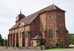 Église en grès rose de trois quarts. Successivement : nef, chevet sans transept et le sommet d'un clocher comtois en tuiles vernissées rouges.