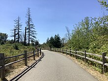 A wide, paved, slightly windy trail flanked by wood fence is in the foreground, with a small metal bridge in the background.