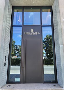 One can see a large black door surrounded by glass panes found on the sides and over the door itself. The whole thing is kind of embedded in the beige walls of the building.