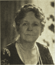 B&W portrait photo of a middle-aged woman with wavy hair in an up-do, wearing a pearl necklace and dark blouse.