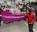 Olena Schewtschenko ist in einer roten Jacke und mit einem Megafon auf einer Demonstration zu sehen. Im Hintergrund sind viele andere Menschen die ein pinkfarbenes Trnsparent halten.
