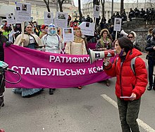 Olena Schewtschenko ist in einer roten Jacke und mit einem Megafon auf einer Demonstration zu sehen. Im Hintergrund sind viele andere Menschen, die ein pinkfarbenes Transparent halten.