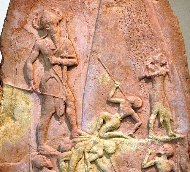 Photographie d'une partie d'une stèle sculptée en bas-relief. On y voit, à gauche, un grand personnage debout, coiffé d'un casque à deux cornes. Il marche vers la droite où se situent quatre petits personnages tombés ou suppliants. Au-dessus de ces-derniers est gravée une inscription en écriture cunéiforme.