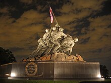 Color photo of the Marine Corps War Memorial, a bronze statue of six Marines raising a U.S. flag attached unto a Japanese pipe atop Mount Suribachi.