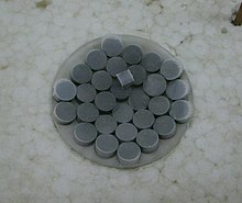 Темно-серые таблетки на стекле. Один кубический кусок того же материала поверх таблеток.