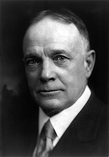 A black-and-white photograph of a balding older man wearing a dark suit and tie