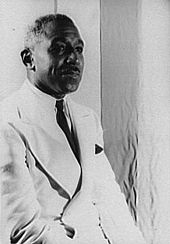 Photo ancienne noir et blanc : trois-quarts face du buste d'un homme noir souriant, les cheveux grisonnants, habillé d'un costume clair et arborant une cravate.