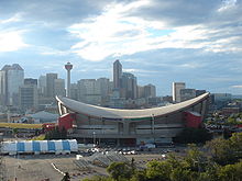 Exterior shot of an indoor arena. The building has a sloped roof in the shape of a reverse hyperbolic paraboloid and a primarily-concrete outer facing with red towers at the corners. Several skyscrapers are visible in the background.