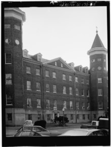 Черно-белая фотография фасадного кирпичного дома с двумя пристроенными к фасаду башнями. На переднем плане видны крыши машин