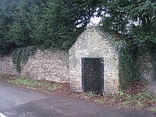 A small stone building with wrought iron gates against a long stone wall. Huge yew trees overhang the wall, and a straggly bit of ivy clings to theside of the well house