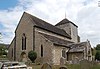 Three-quarter view of a church with flint walls and tiled roofs. A short tower with a pyramidal roof and two small windows rises slightly right of centre. To the left, a longer section with a high roof, with projections with lower rooflines and two low windows to both sides. A porch with an arched doorway extends from the far right side.