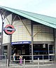 A dark grey building with a green roof and a rectangular, blue sign reading "HOUNSLOW EAST STATION" in white letters all under a blue sky