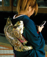 A young woman wearing a dark blue furisode; her obi, which is gold and covered in roundels, is tied like a bow with an oversized middle portion, with two small 'wings' poking out of each side at the top.