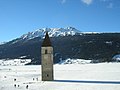Cerkwiny torm pśi Reschensee (Lago di Resia) pód sněgakowańskego stronom Elferspitze (Cima Undici) a Schöneben (Belpiano).