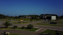 Uma foto dos arredores das instalações da Universidade Federal do Rio Grande. Está de manhã, e o céu está azul. Há muitas áreas gramadas, com plantas menores rasteiras, e algumas pistas. O prédio da universidade está ao longe, no horizonte, e tem paredes acinzentadas.