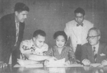 Фотография молодой женщины, смотрящей на газету, в окружении четырех мужчин.