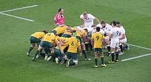 Deux équipes de rugby se préparant à disputer une mêlée, avec au premier plan les deux demis de mêlée, l'arbitre positionné sur le côté opposé.