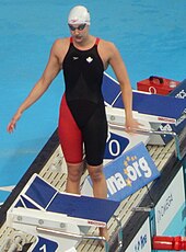 Chantal van Landeghem steht neben dem Startblock und trägt einen dunkelblauen Badeanzug mit auf einer Seite rotem Beinansatz. Auf dem Badeanzug ist ein weißes Ahornblatt erkennbar. Sie hat eine weiße Badekappe auf und ihre Schwimmbrille verdeckt ihre Augen.